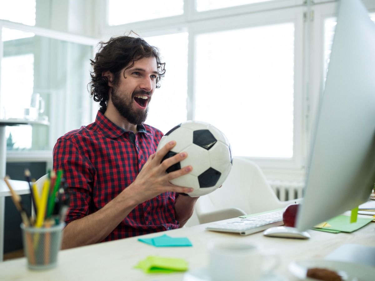Um homem segurando uma bola de futebol nas mãos enquanto sorri e olha para a tela do computador - Discuta os próximos lançamentos de jogos com seu público do YouTube - Imagem