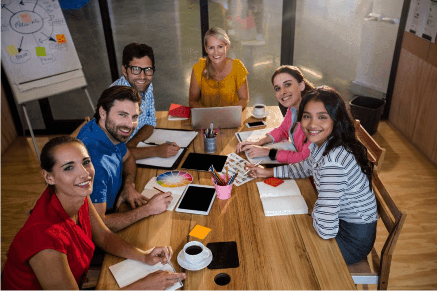 Des collègues assis à un bureau et souriant devant la caméra – Présenter votre culture de travail sur YouTube est un bon moyen de promouvoir votre marque – Image