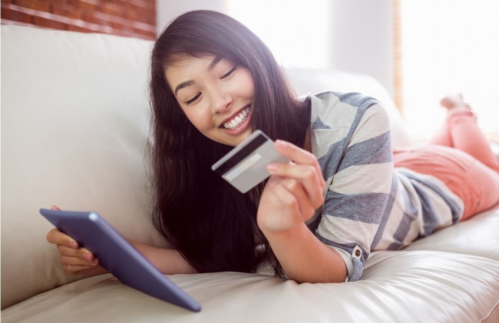 A young woman with a phone and a credit card in her hand lies on the sofa and smiles - Customer loyalty programs are a great way to engage your customers - Image