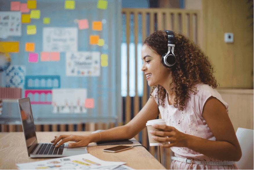 A young woman wearing headphones holds a cup of coffee and uses a laptop - Important tips for vision board design - Image