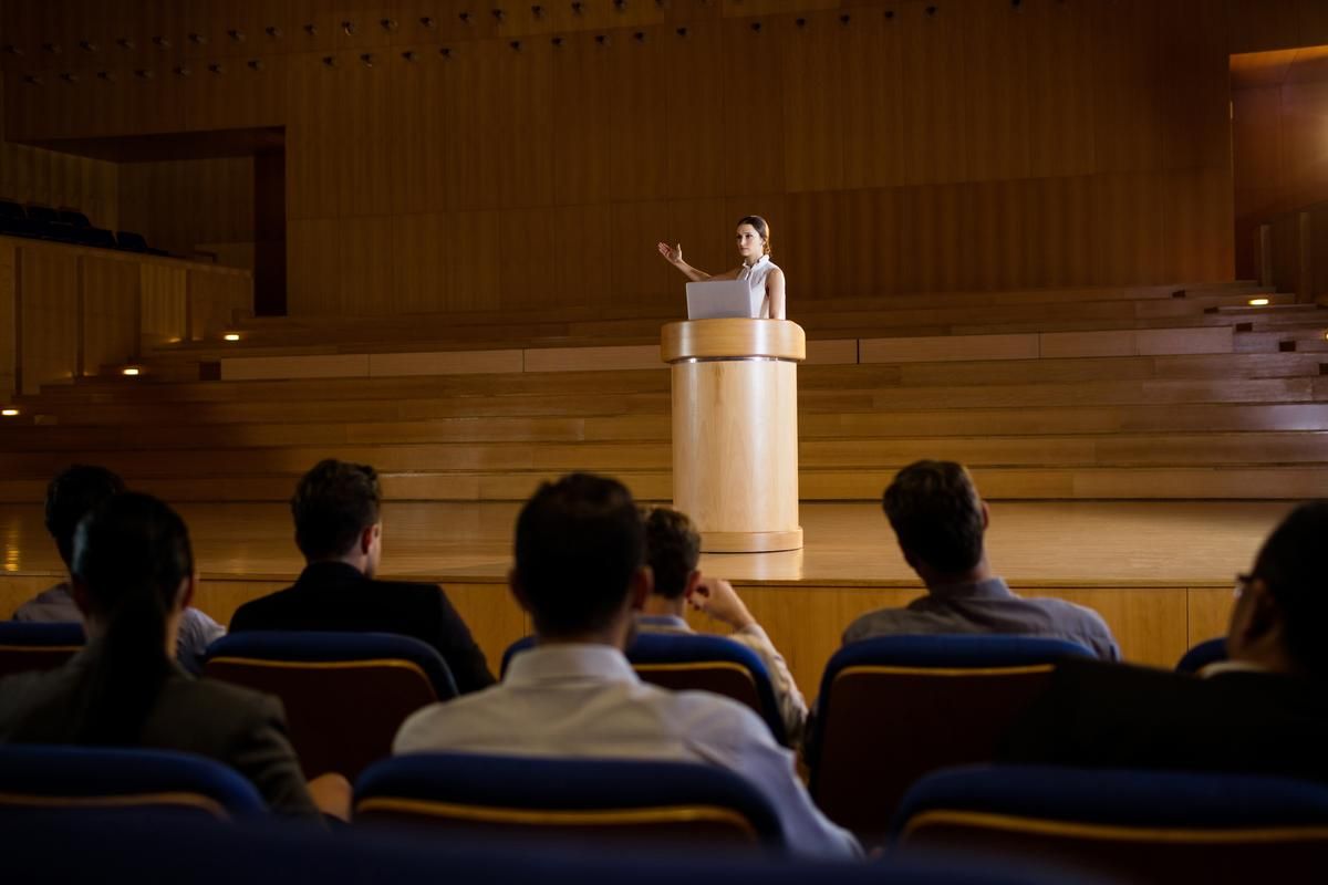 A woman speaks at a lectern in front of an audience - How to get more views on YouTube: The ultimate guide - Image