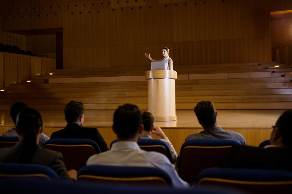 A woman speaks at a lectern in front of an audience - How to get more views on YouTube: The ultimate guide - Image