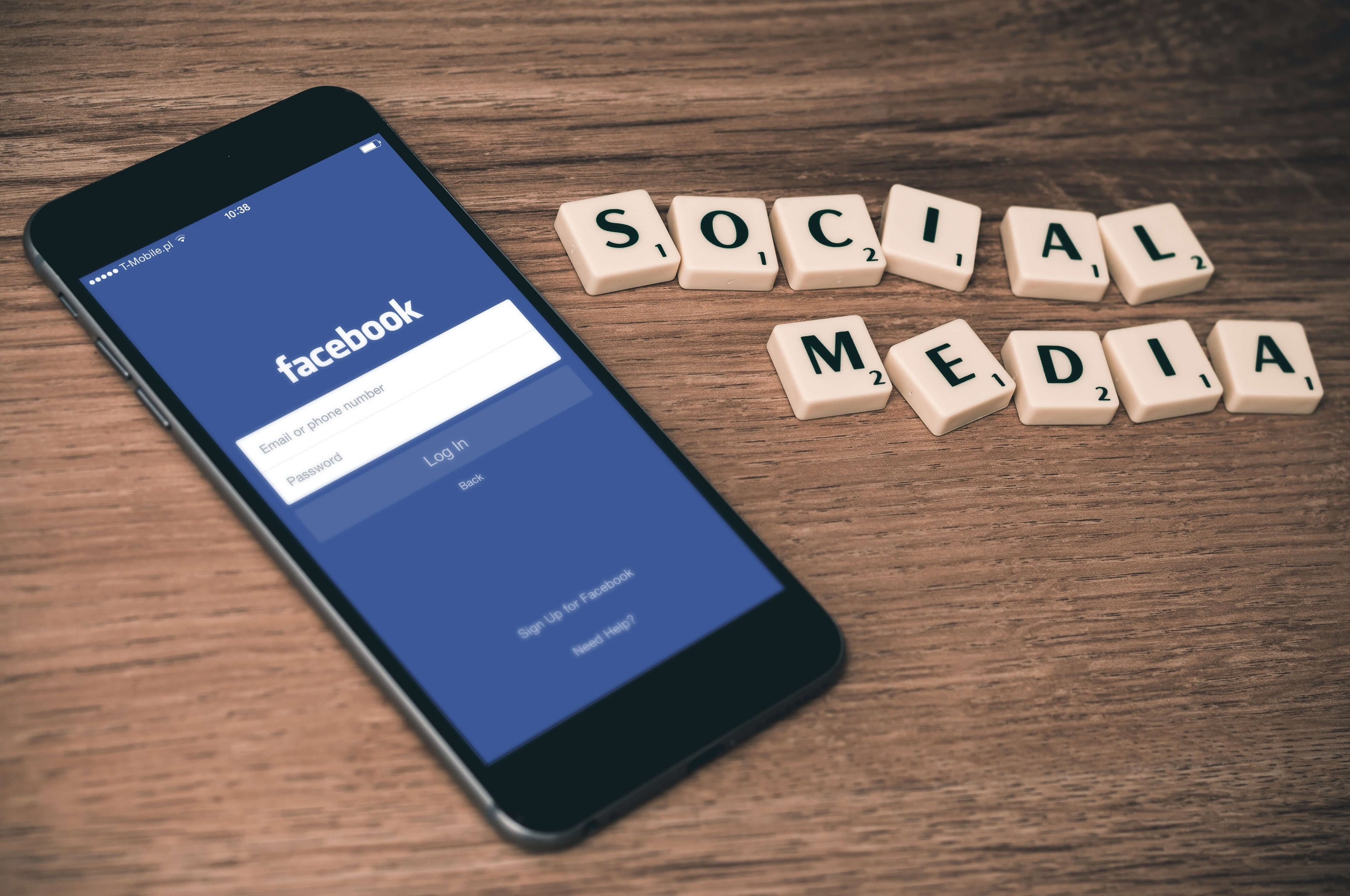 The image shows a smartphone with the Facebook login page open and the text 'Social Media' made up of Scrabble letters next to it on a wooden table - Facebook ads: A step-by-step guide for beginners - Image