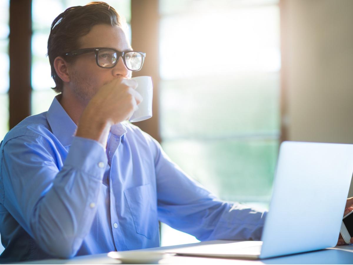 A male executive with glasses drinks coffee while working on a laptop; Facebook profile picture - Tips for choosing the best profile photo for different social networks - Image